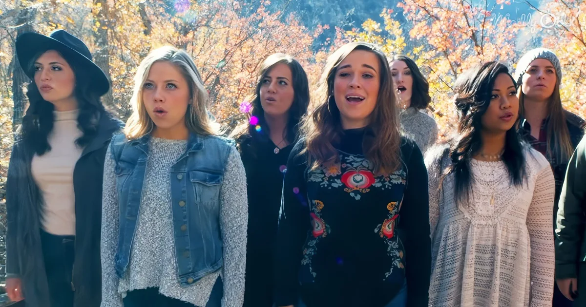 A group of six women, dressed in casual attire, stand together outdoors with autumn foliage in the background.