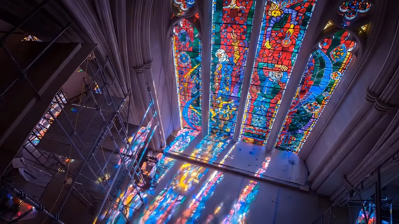 Stained glass windows with vibrant colors and religious figures are illuminated, casting colorful light inside a cathedral.