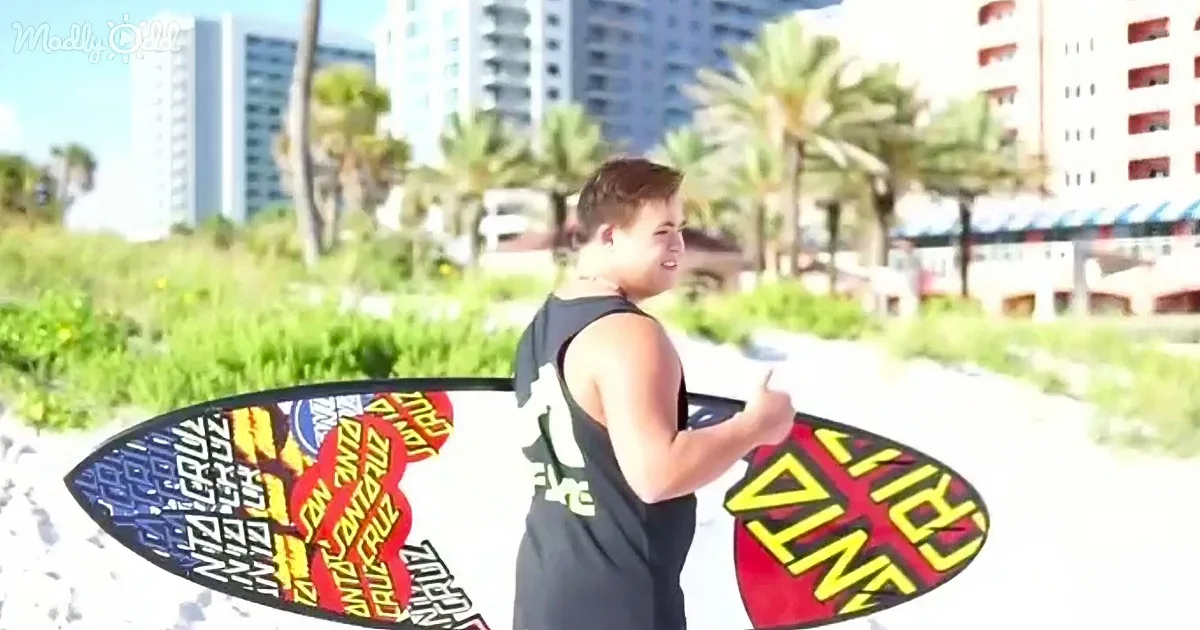 A person in a black tank top holds a colorful surfboard, giving a thumbs-up near a beach with palm trees and buildings.