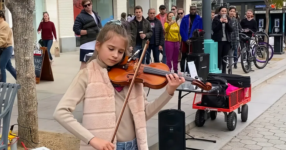 A young girl plays the violin on a sidewalk, surrounded by onlookers and her violin case.