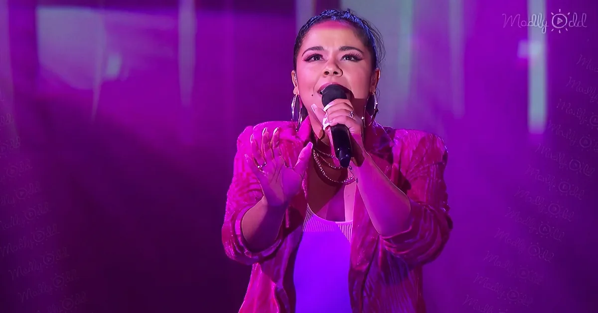 A person in a pink outfit sings into a microphone on a stage with purple lighting.