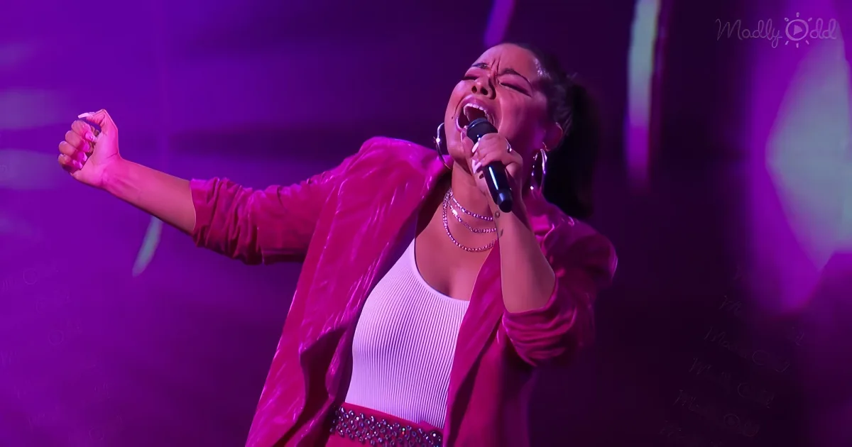 A woman in a pink outfit sings passionately on stage with a microphone, surrounded by purple lighting.
