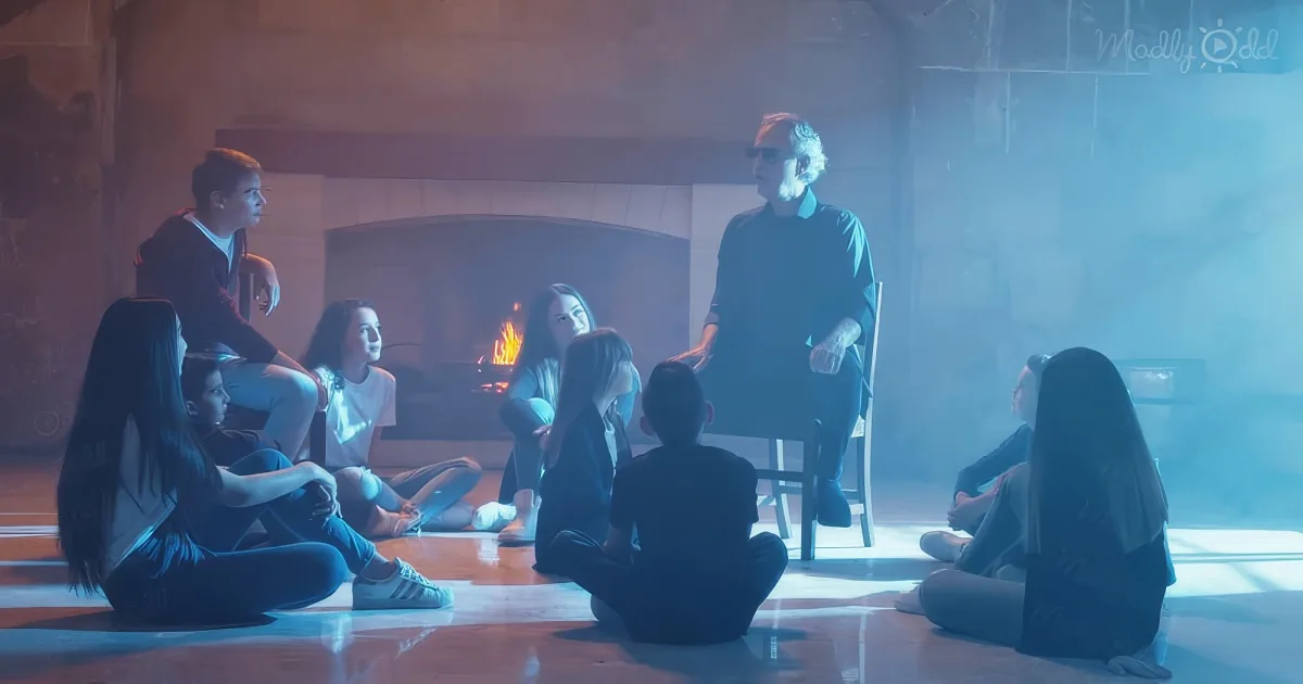 A man stands in front of a group of seated people in a dimly lit room with a fireplace.
