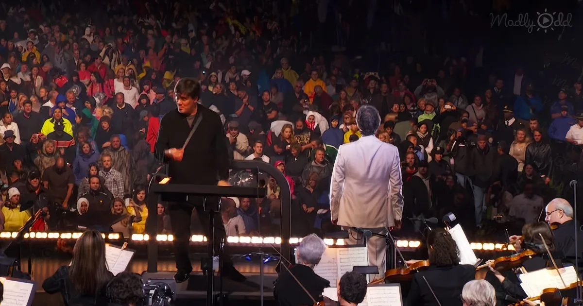 A man in a black jacket stands on stage, facing a conductor and an orchestra in front of a large audience.