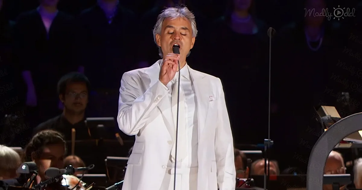 A man in a white suit sings into a microphone on stage with an orchestra behind him.