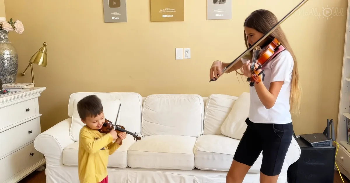 A girl plays violin on a couch, while a boy sits behind her, both in a cozy living room setting.