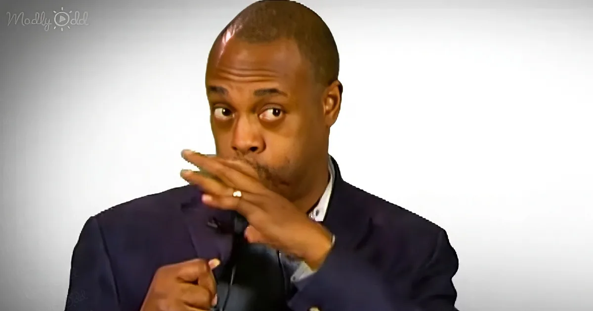 A man in a suit holds a microphone, hand near mouth, appearing to speak or react.