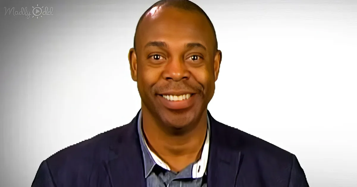 A smiling man in a suit and tie, with a white background and a logo in the top left corner.