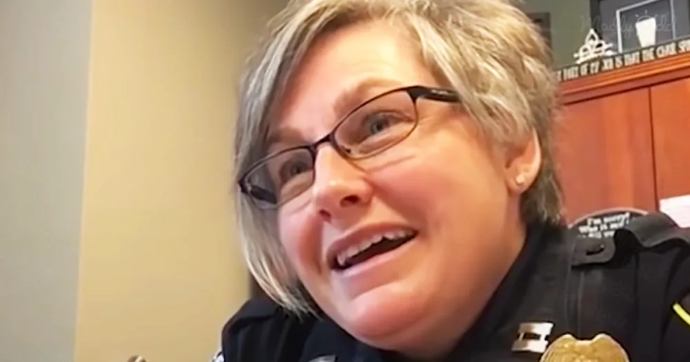A woman in a police uniform is smiling and wearing glasses, with a badge visible on her uniform.