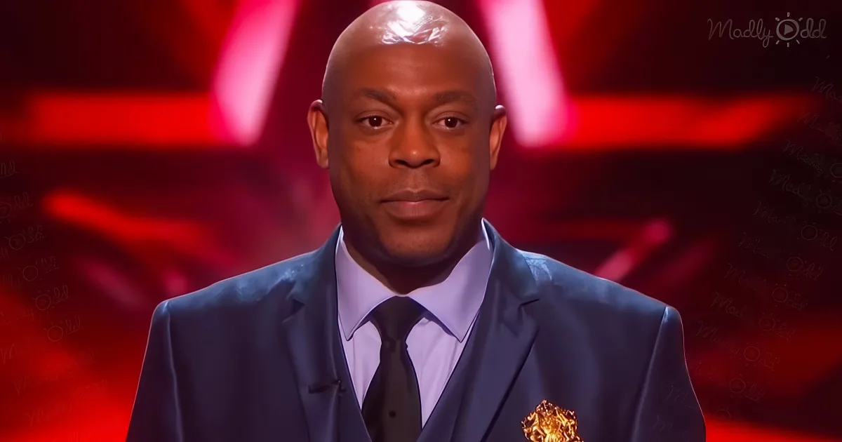 A man in a suit holds an award, smiling, with a red background and "Madly Odd" watermark.