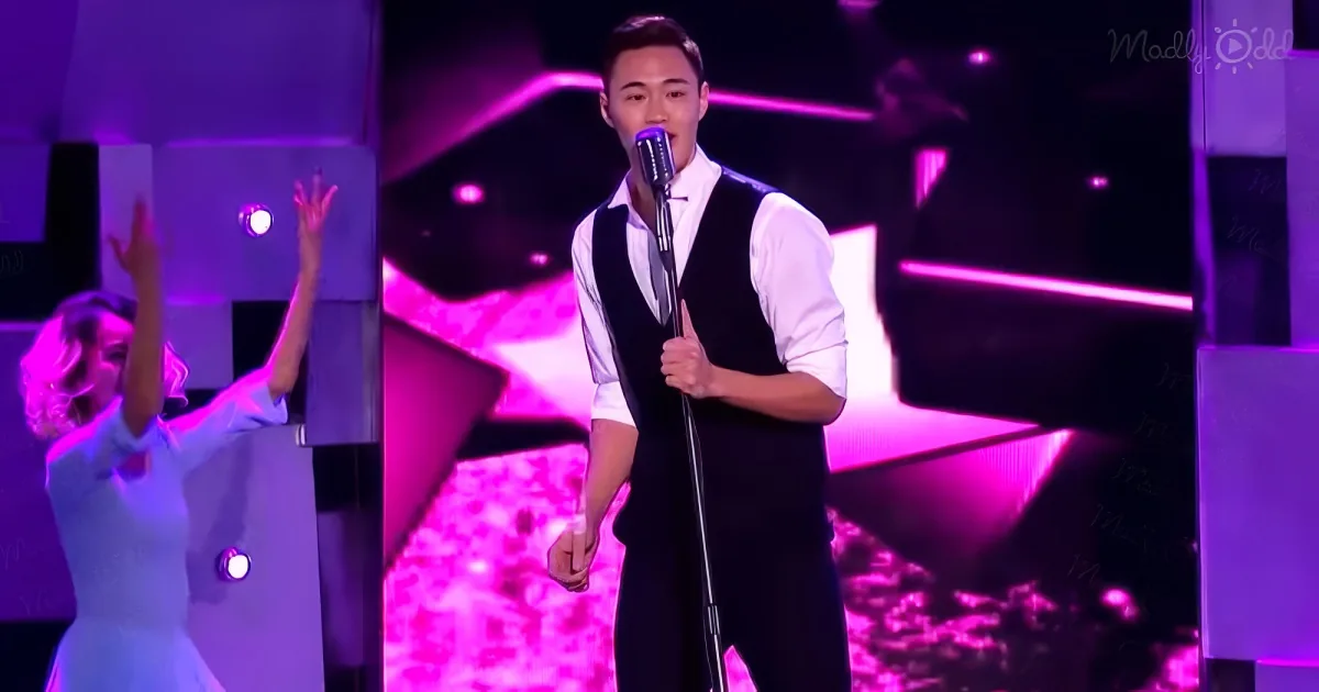 A man in a vest sings into a microphone on stage with vibrant purple lighting.