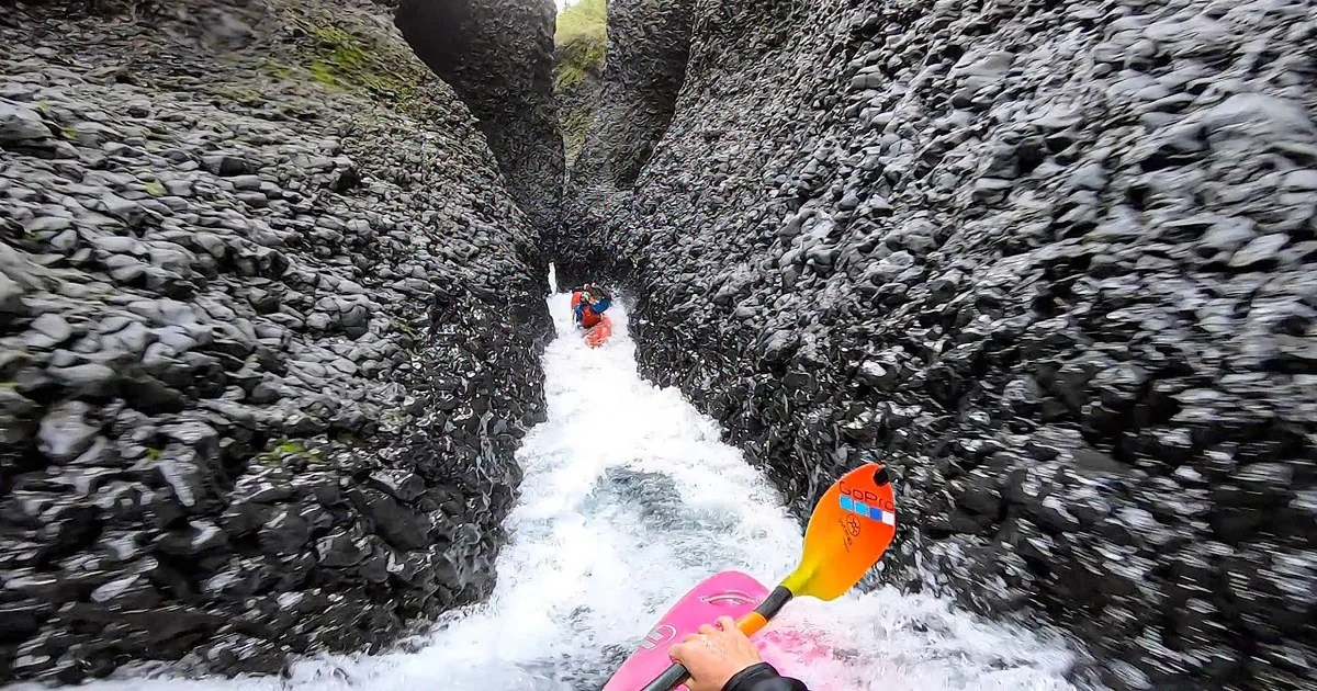 A kayaker in a red life jacket navigates a narrow, rocky river passage with a pink kayak and paddle.