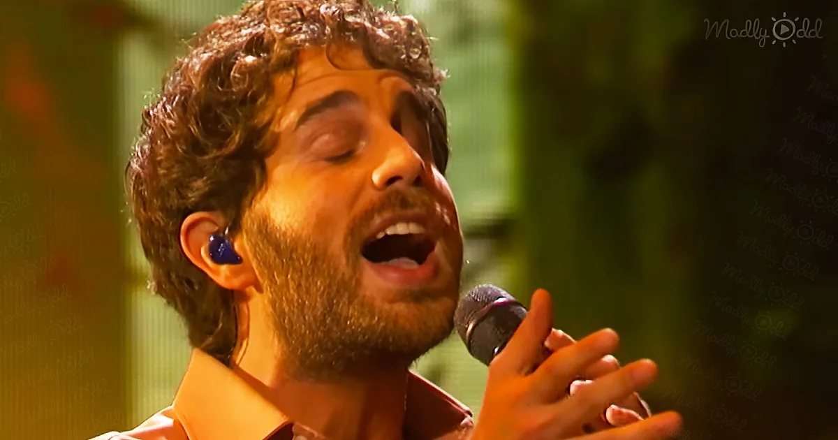 A man with curly hair and a beard sings passionately into a microphone, wearing an earbud.