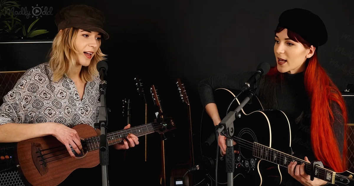 Two women play acoustic guitars and sing into microphones, one with blonde hair and a hat, the other with red hair and a...