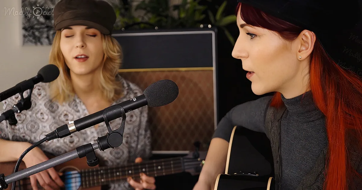 A woman with red hair sings into a microphone while playing guitar, with another person in the background.