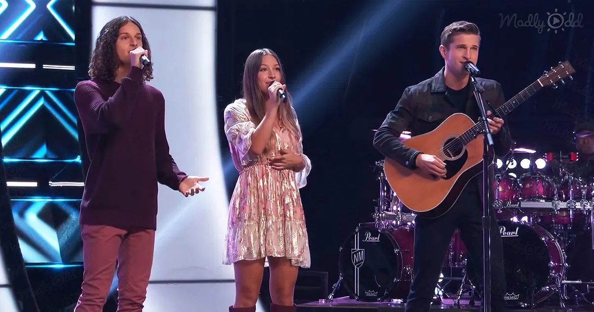 Three performers sing on stage, one with a guitar, another with a microphone, and the third holding a mic.