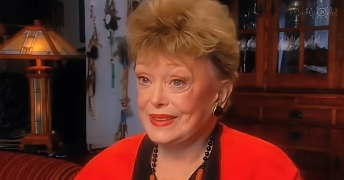 A woman in a red outfit is sitting, wearing a necklace, with a lamp and wooden furniture in the background.
