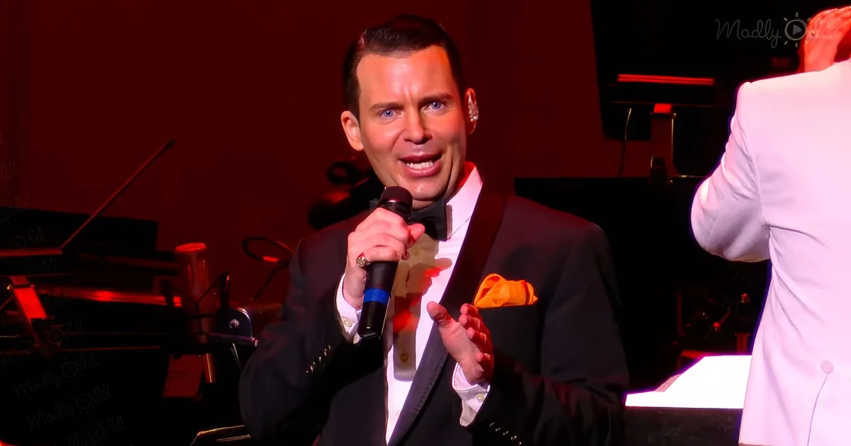 A man in a tuxedo sings into a microphone on stage, with a piano and musicians in the background.