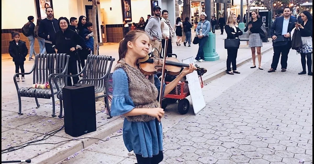 A young girl in a blue dress and fur vest plays the violin on a city sidewalk, surrounded by onlookers.