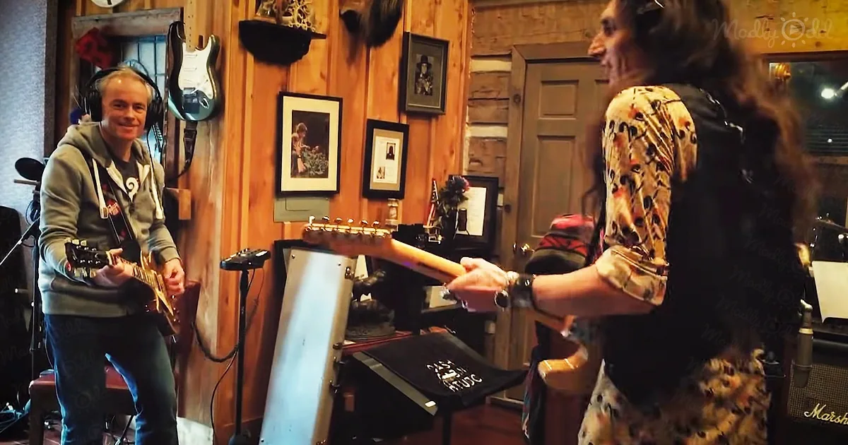 A man in a hoodie plays guitar while another person in a floral shirt holds a guitar neck.