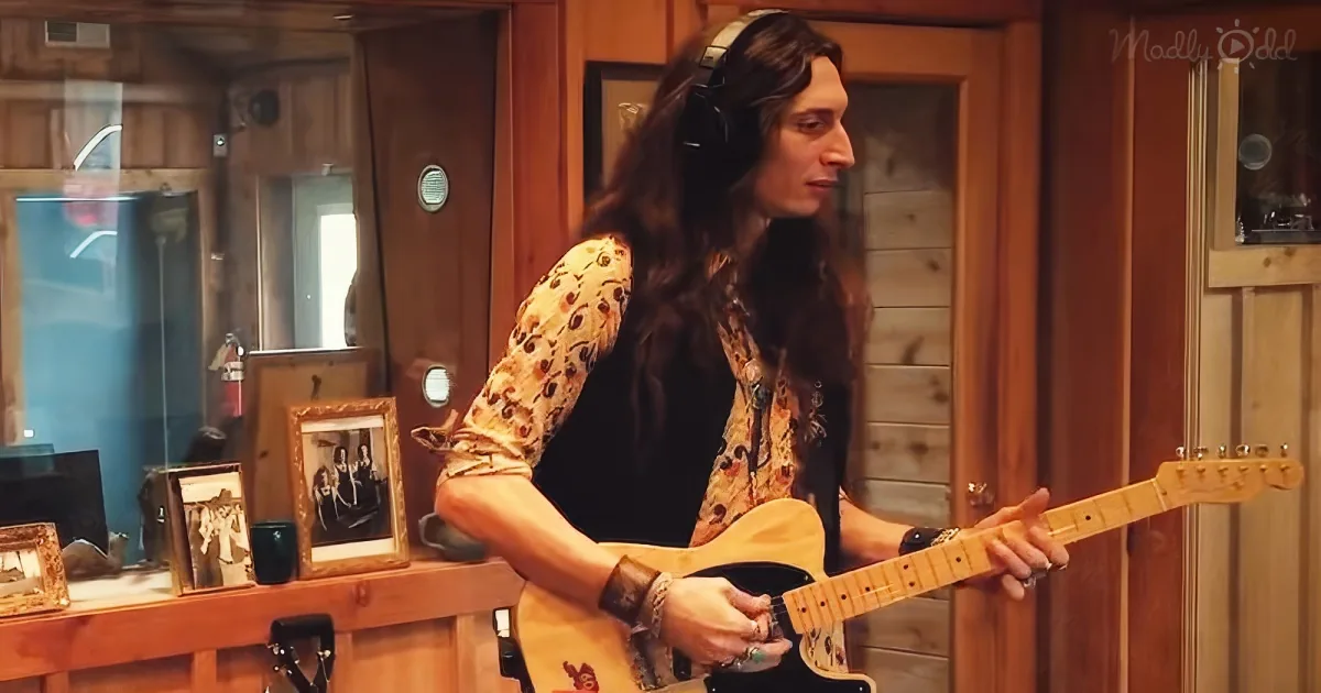 A person with long hair and headphones plays a guitar in a cozy, wood-paneled room.