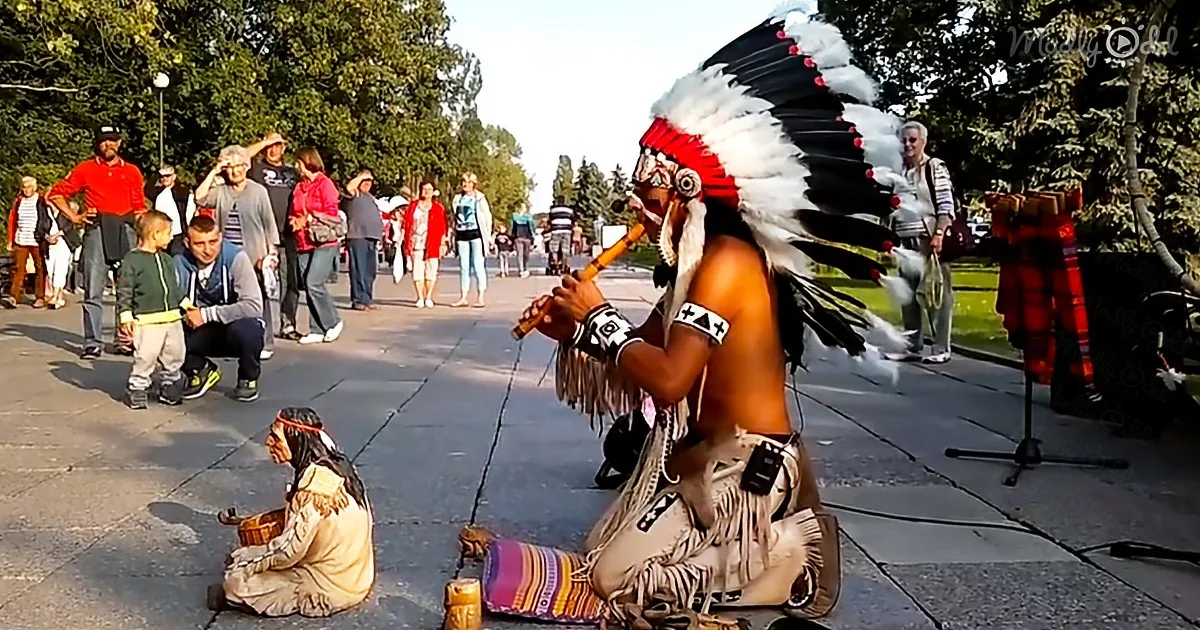 A man in traditional Native American attire plays a flute while another person in similar attire sits nearby, surrounded...