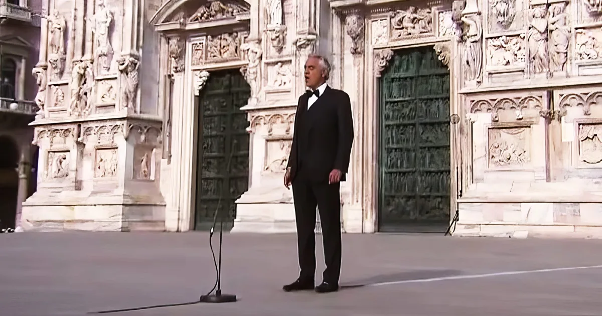 A man in a tuxedo stands on steps in front of ornate doors, likely performing or singing.