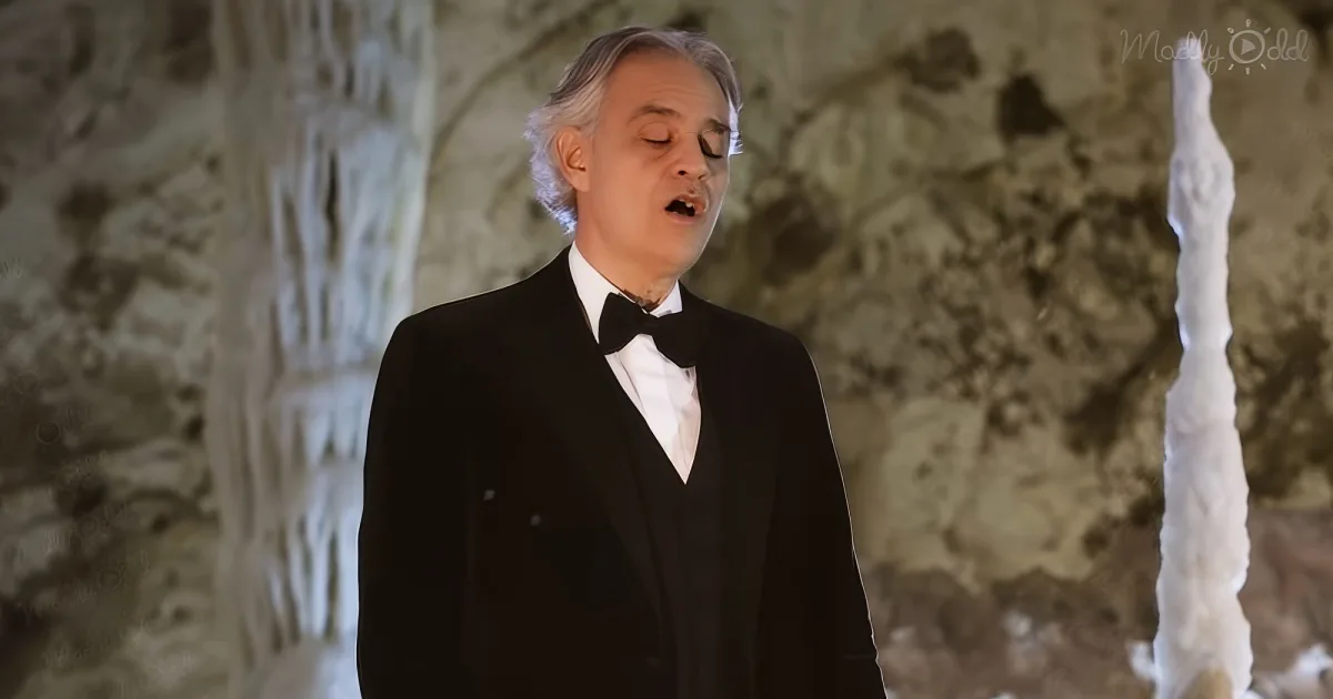 A man in a tuxedo sings passionately in front of a stone wall backdrop.