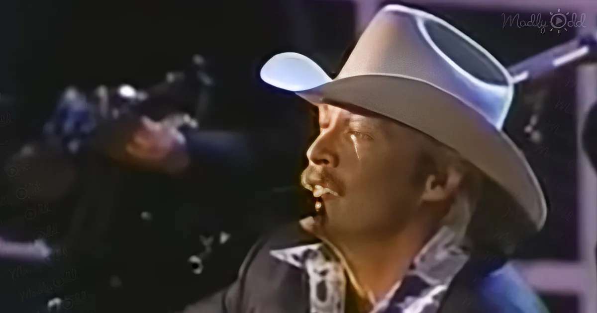 A man in a cowboy hat is singing into a microphone, holding a guitar, with a dark background.