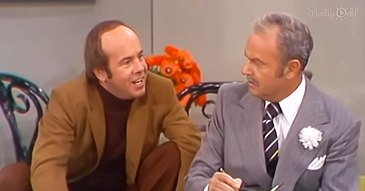 A man in a brown suit is smiling while another in a gray suit listens, both seated indoors.