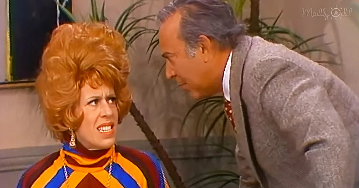 A woman with a large red wig and colorful outfit is speaking to a man in a suit, both in a room with plants.