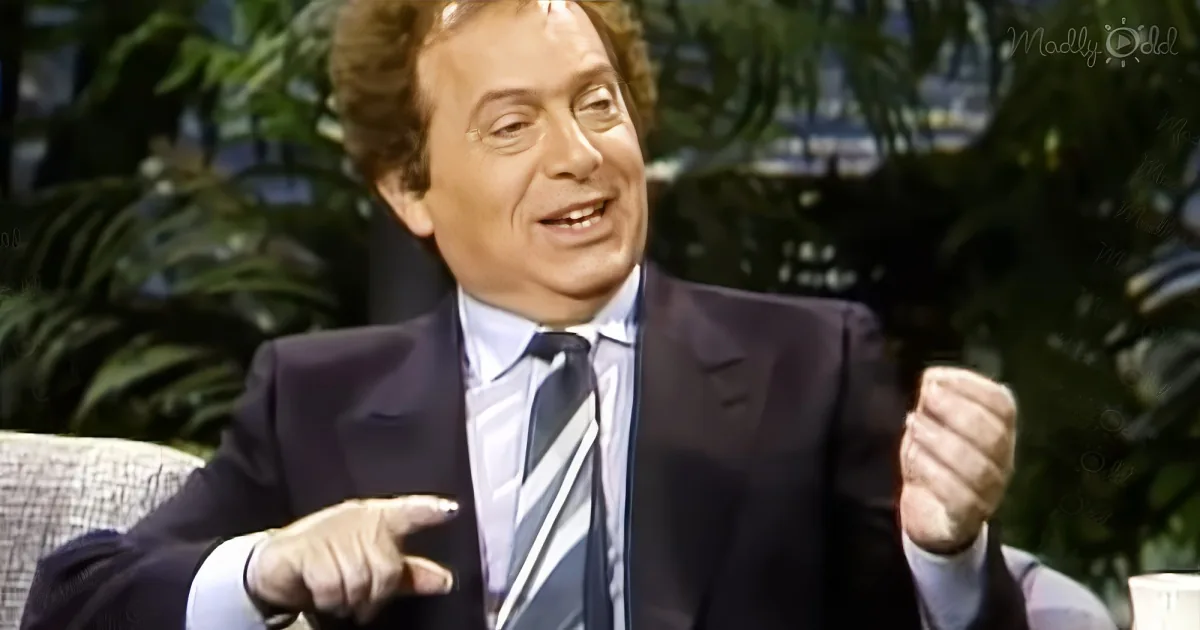 A man in a suit and tie is gesturing while sitting on a couch, with a plant in the background.