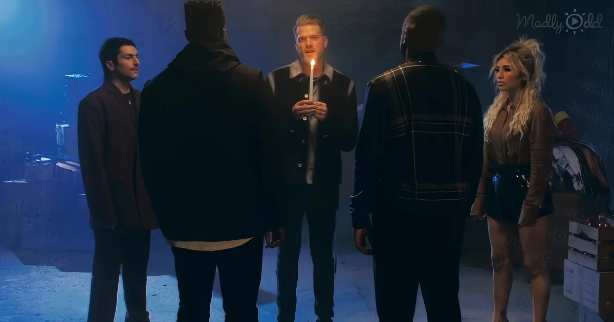 A man holds a lit candle, surrounded by four others in a dimly lit room.