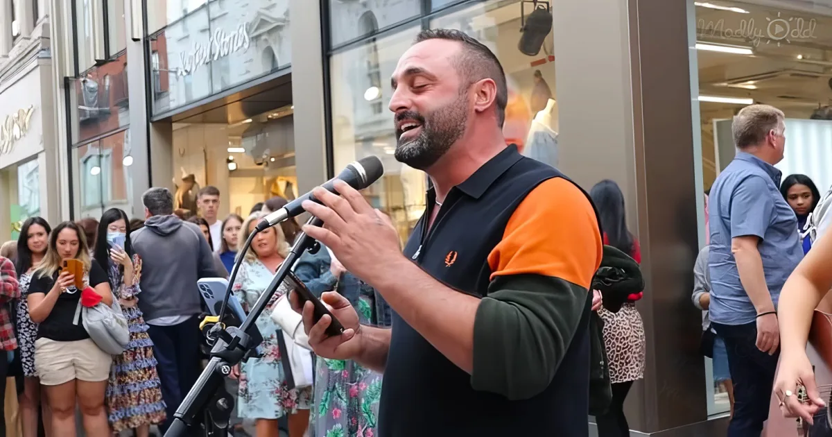 A man in an orange and black shirt sings into a microphone while a crowd watches in a bustling urban setting.