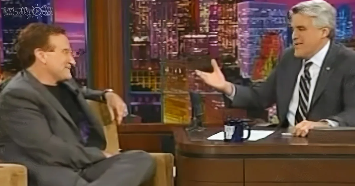 A man in a suit gestures while seated at a desk, engaging in conversation with another man in a black shirt.