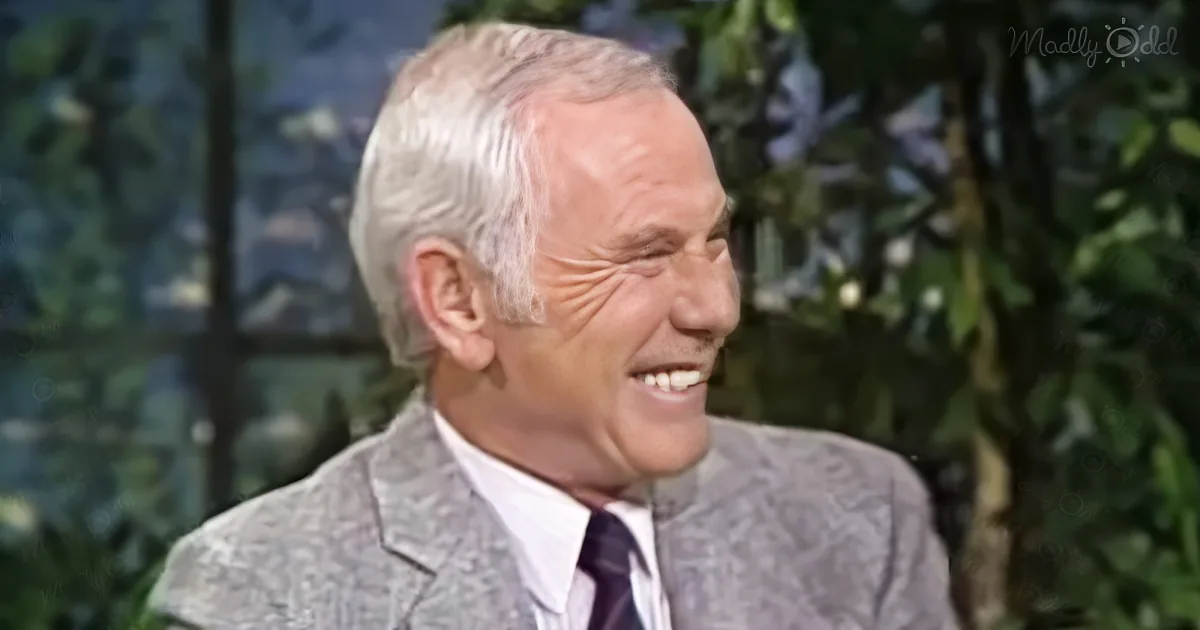An older man with white hair, wearing a suit and tie, smiles while seated in front of a blurred green background.