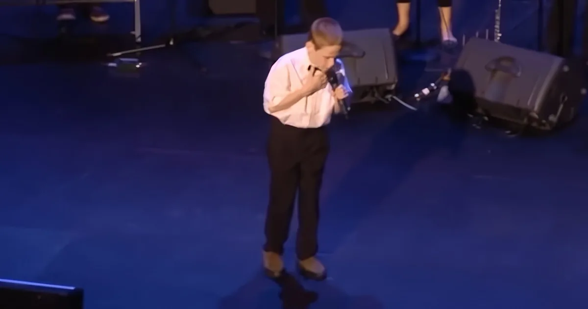 A young boy in a white shirt and black pants holds a microphone on a stage with blue lighting.