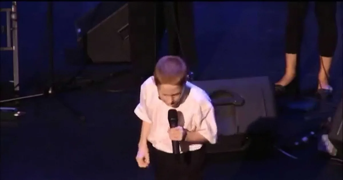 A young person in a white shirt holds a microphone, standing on a stage with a dark background.