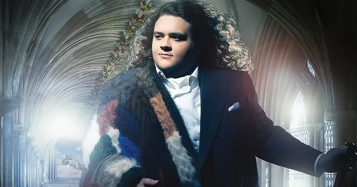 A man with long, curly hair in a suit and fur coat stands in a grand, arched hallway with sunlight streaming through.
