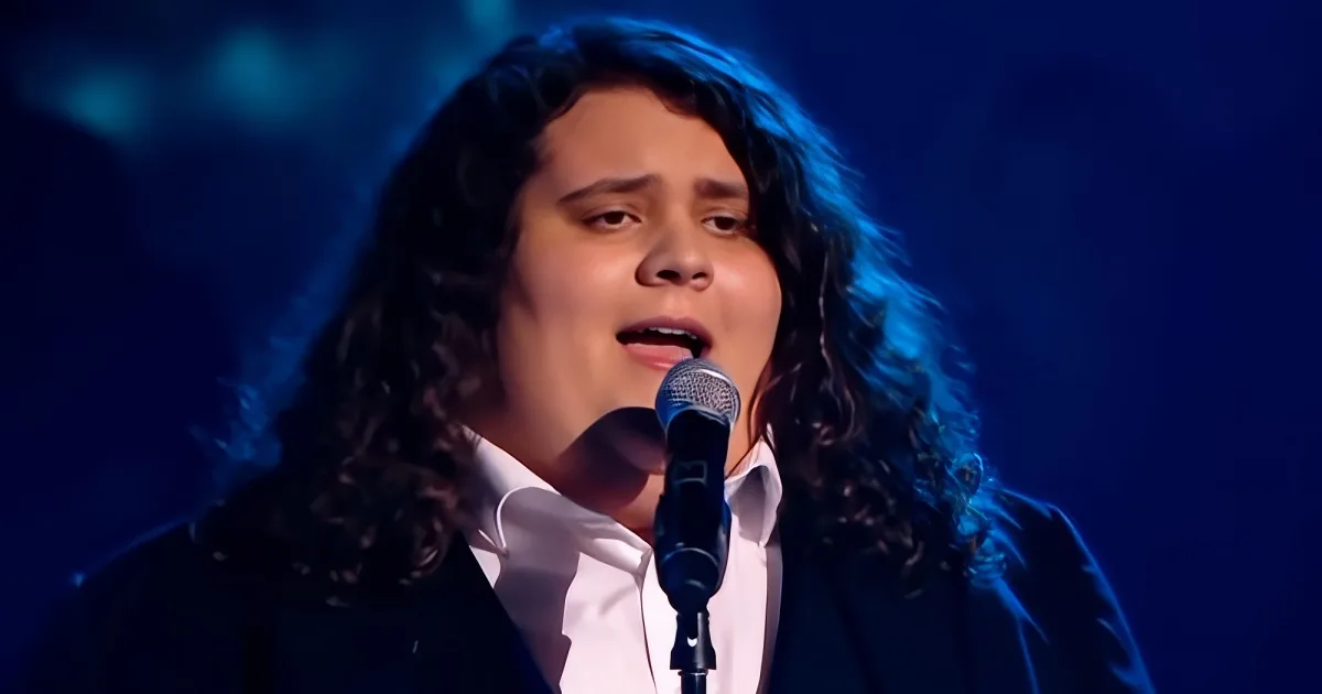 A person with curly hair sings into a microphone on stage under blue lighting.