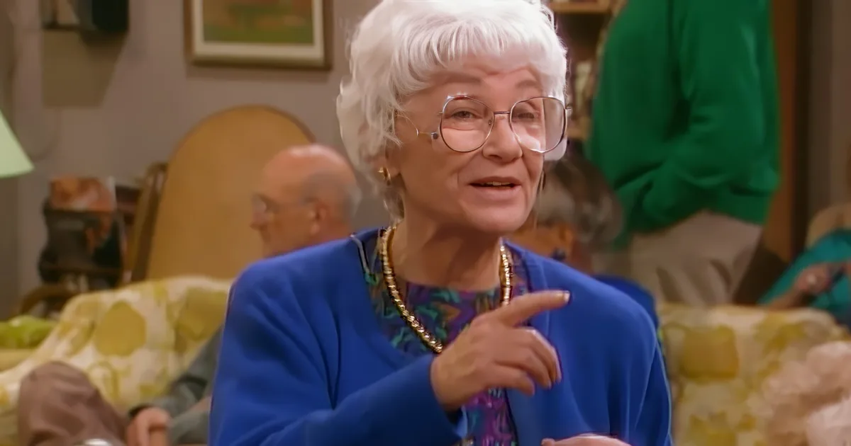 An elderly woman with white hair and glasses gestures animatedly while wearing a blue jacket and gold necklace.