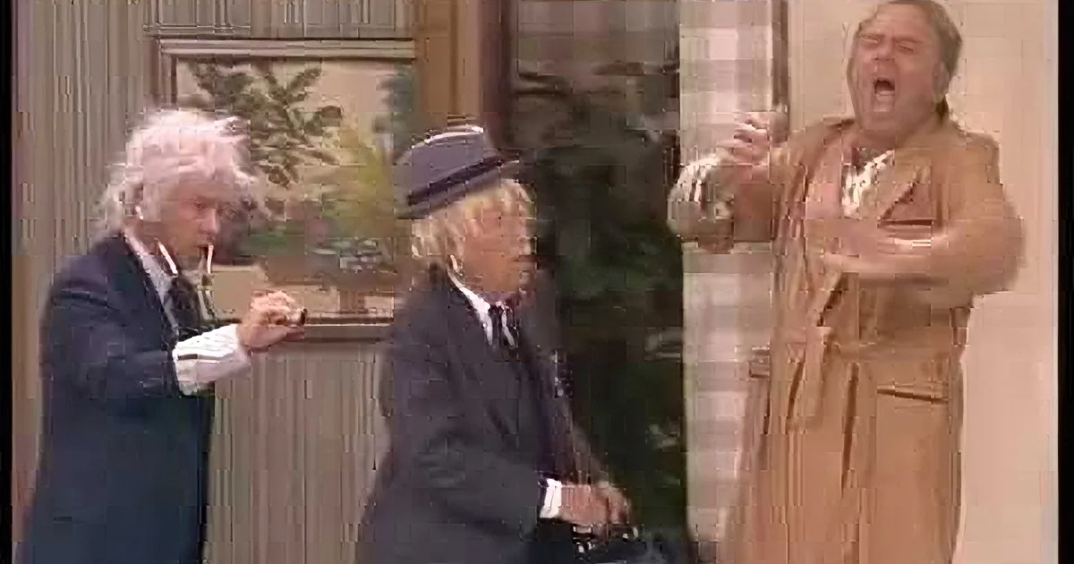 A man in a suit holds a flute while another man in a brown suit lies on a couch, both in a room with a checkered wall and...