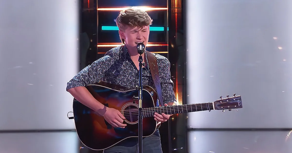 A young man in a floral shirt plays an acoustic guitar and sings into a microphone on stage.