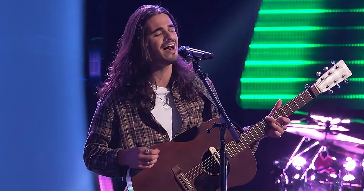 A man with long hair and a plaid shirt sings passionately while playing an acoustic guitar on stage.