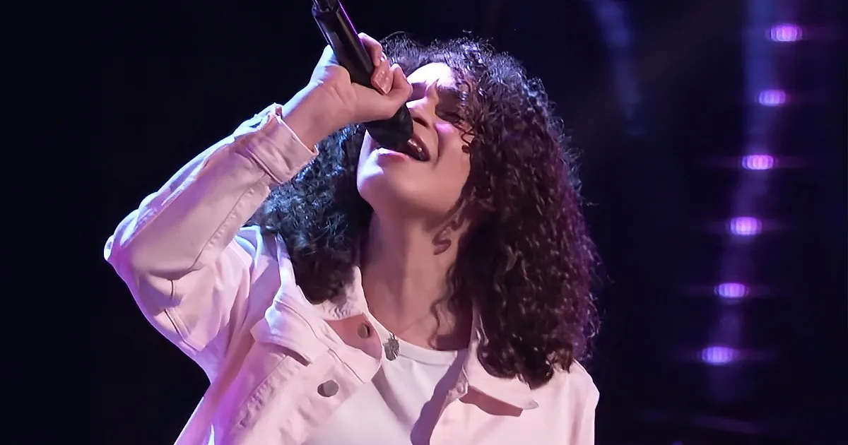A person with curly hair, wearing a light pink jacket, sings into a microphone on stage with purple lighting.