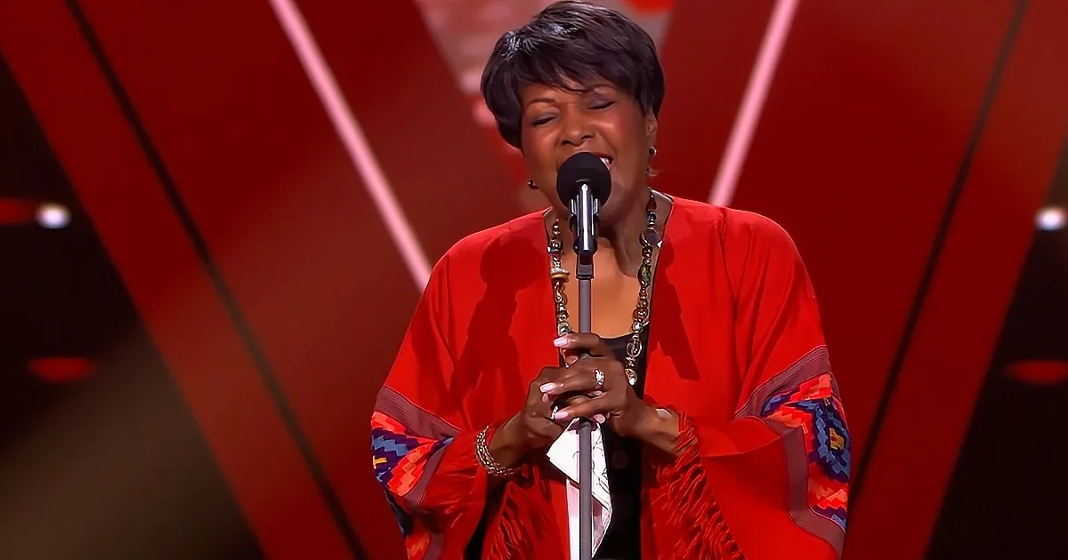 A woman in a red outfit sings into a microphone on a stage with red and black background.