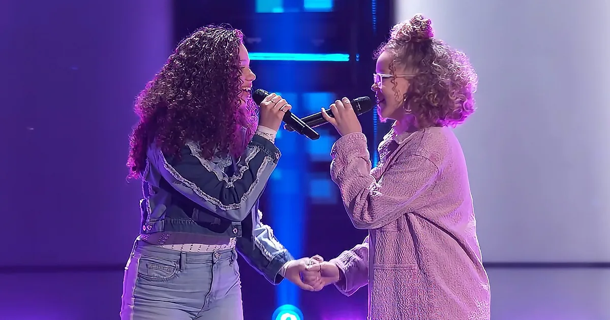 Two women sing together on stage, holding microphones and holding hands, illuminated by purple and blue lights.