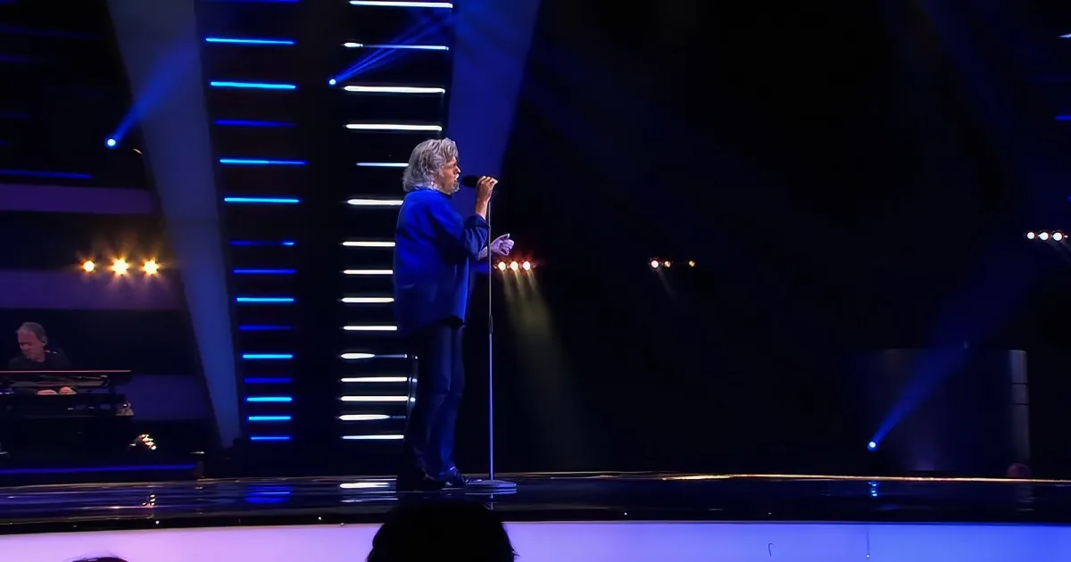 A man in a blue suit stands on stage, holding a microphone, with blue and white lights illuminating the background.