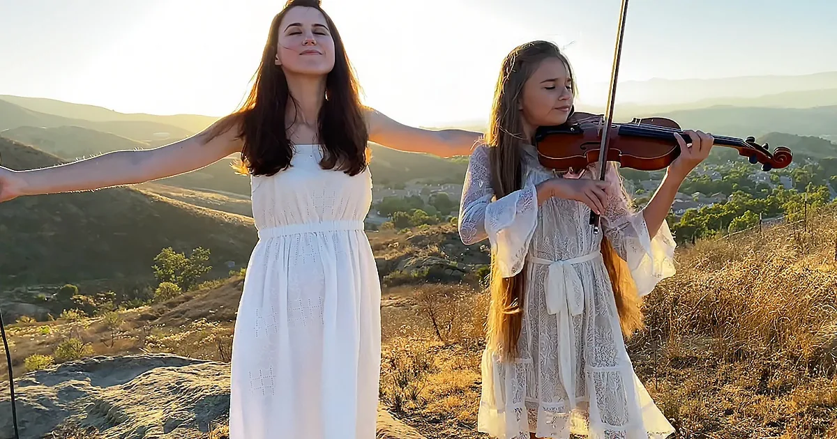 A woman in white dress stands with arms outstretched, while a girl in a white dress plays the violin on a hillside.