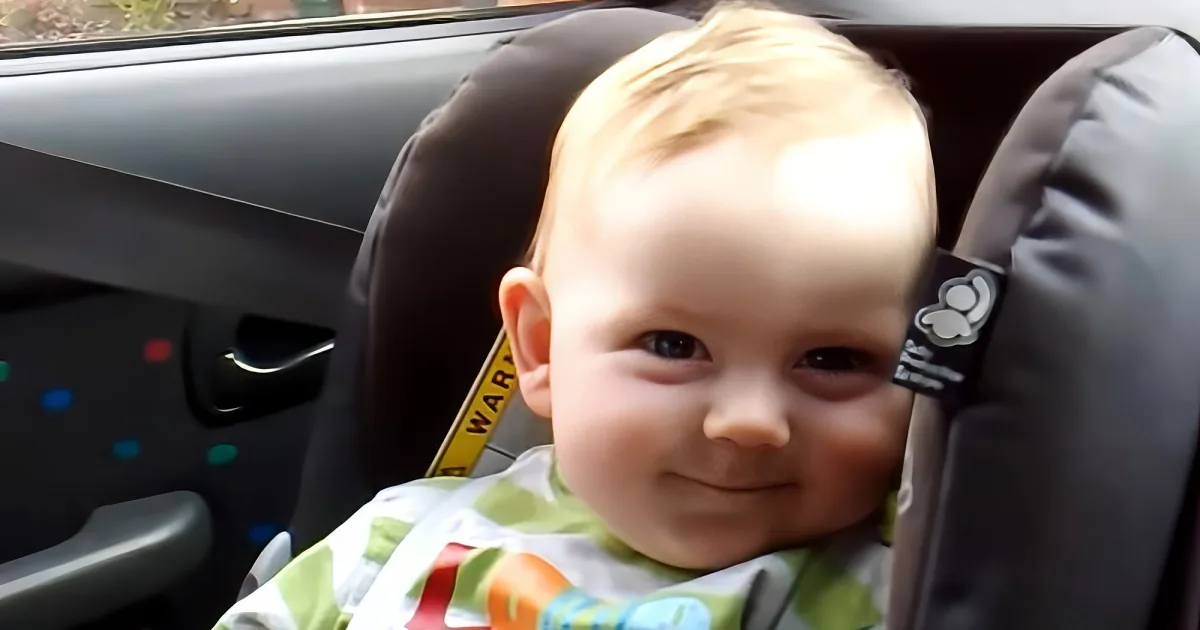 A baby in a car seat, wearing a green and white outfit, with a seatbelt visible.